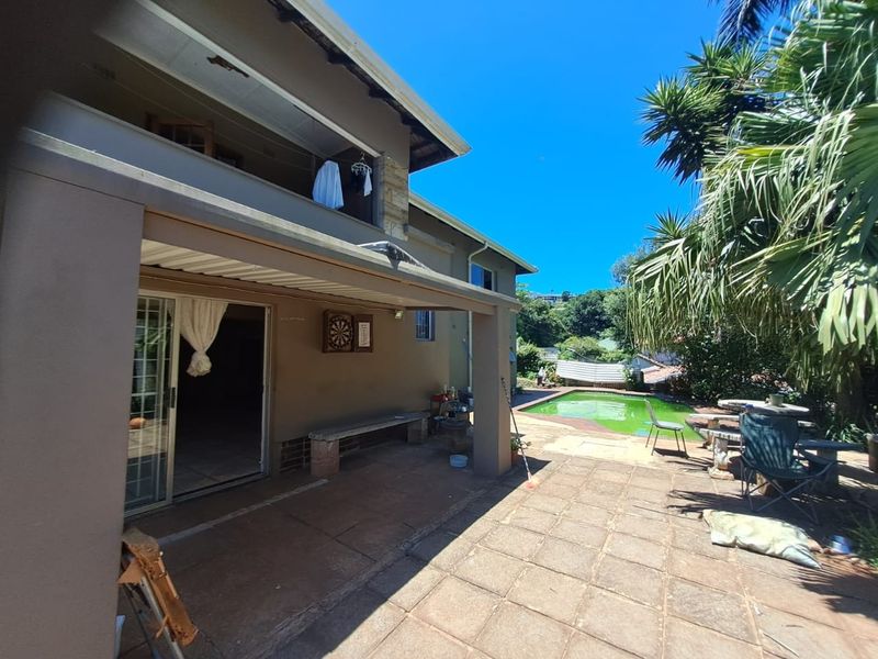 Rear exterior view featuring a covered patio, paved terrace, and a swimming pool, with an upper-level balcony overlooking the garden area.