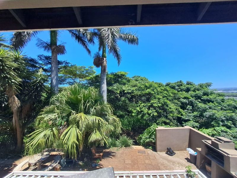 An elevated outdoor patio area featuring a built-in braai, paved flooring, and views over the lush garden and surrounding landscape.