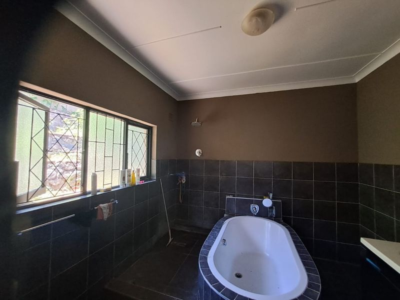 A functional bathroom featuring a built-in bathtub, wall-mounted shower head, and dark tiled wall finishes with security-barred windows.