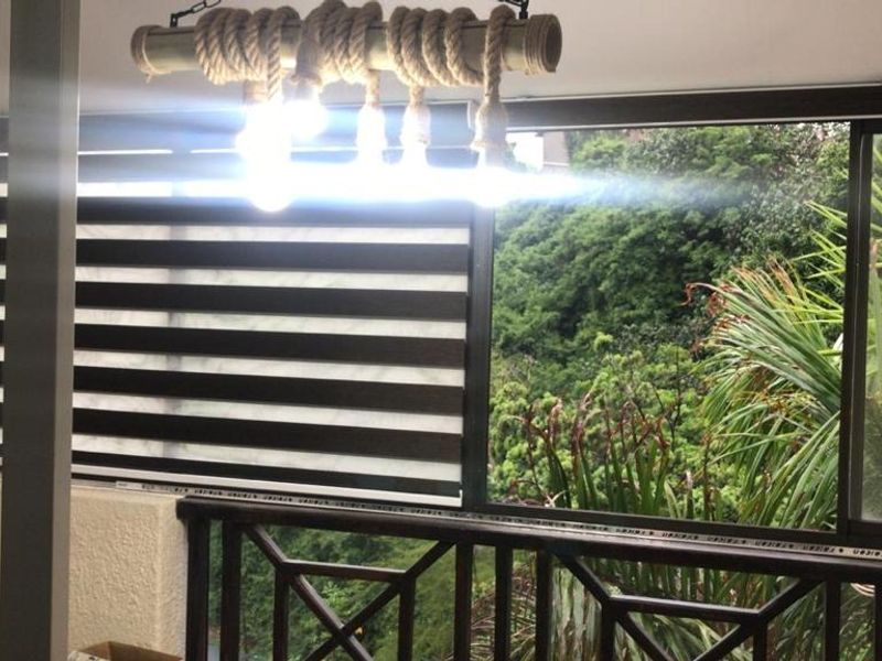 A covered balcony featuring sliding glass doors, modern zebra blinds, and a decorative rope pendant light, overlooking lush greenery.