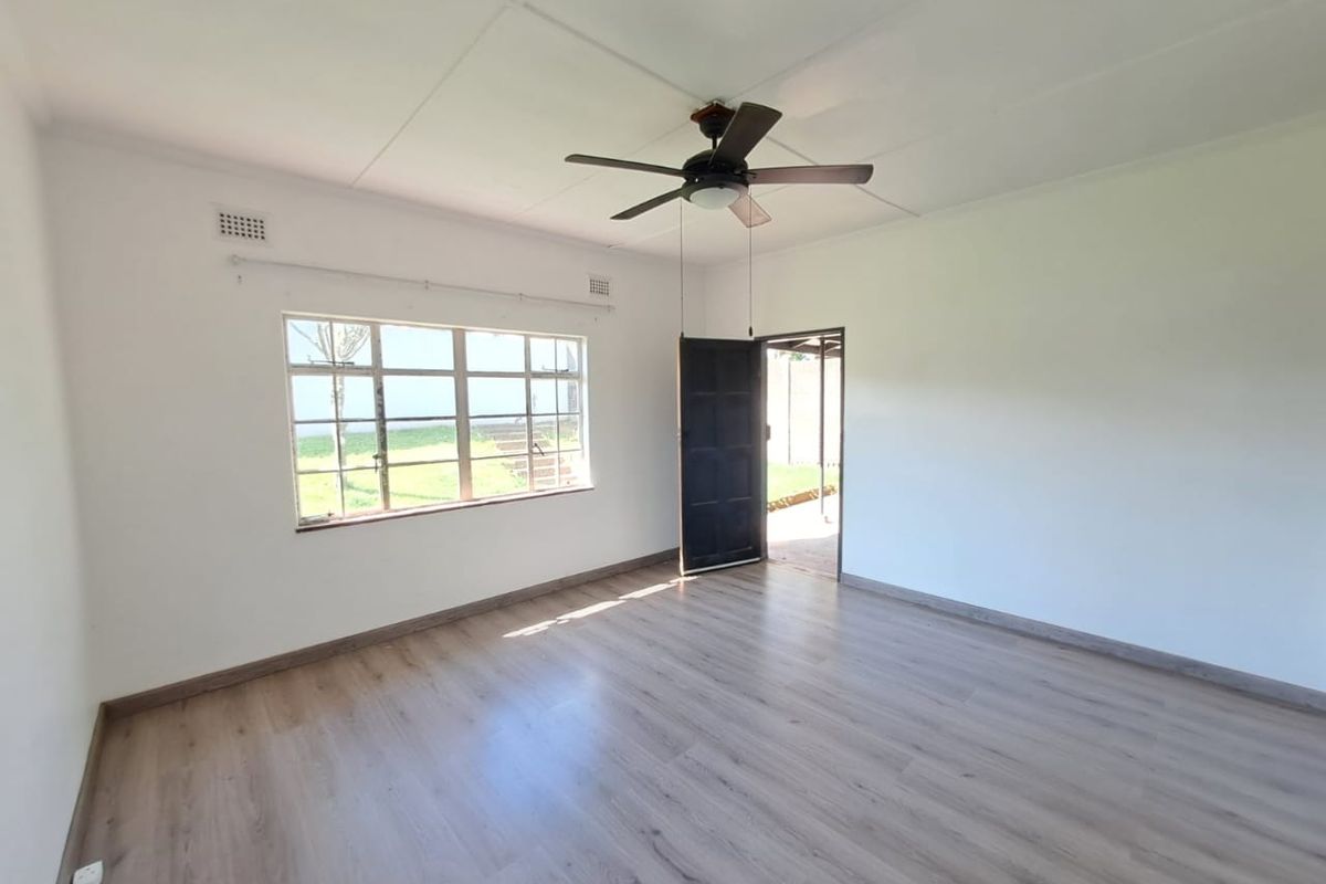 A bright, empty living room featuring modern laminate flooring, a ceiling fan, and large windows providing natural light.