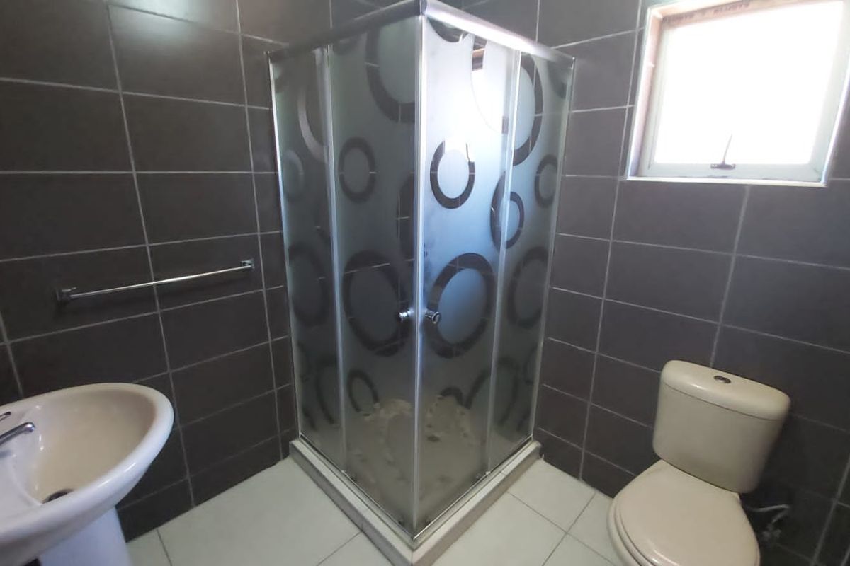 A functional bathroom featuring a corner shower enclosure, pedestal basin, and toilet with dark wall tiling and light floor tiles.