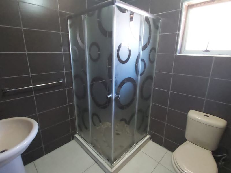 A functional bathroom featuring a corner shower enclosure, pedestal basin, and toilet with dark wall tiling and light floor tiles.