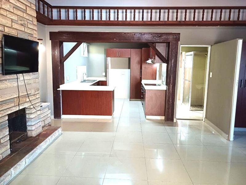 Open plan living area featuring a stone fireplace, timber beam accents, and a functional kitchen with built-in cabinetry and tiled flooring.