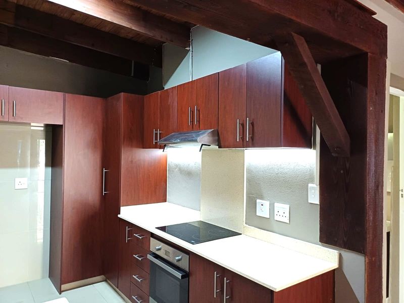 A modern kitchen featuring dark wood cabinetry, stone countertops, integrated electric hob and oven, and exposed timber ceiling beams.