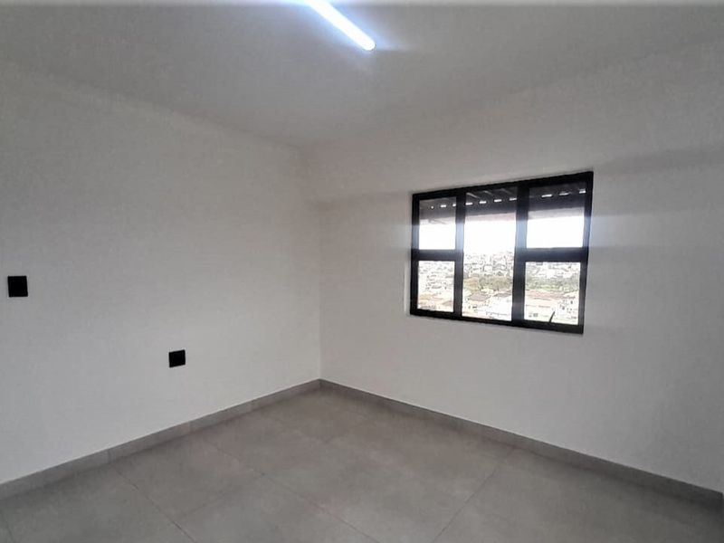 A bright, empty bedroom featuring neutral tiled flooring, white walls, and a black-framed window with an exterior view.