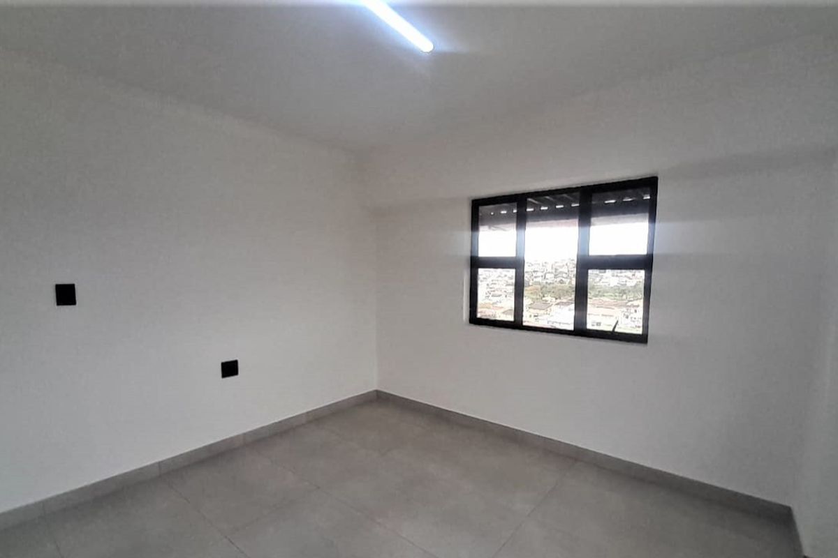 A bright, empty bedroom featuring neutral tiled flooring, white walls, and a black-framed window with an exterior view.