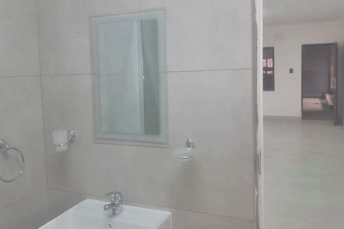 A clean, modern bathroom featuring tiled walls and flooring, a wall-mounted basin with chrome fixtures, and a mirror, leading into an adjacent living area.
