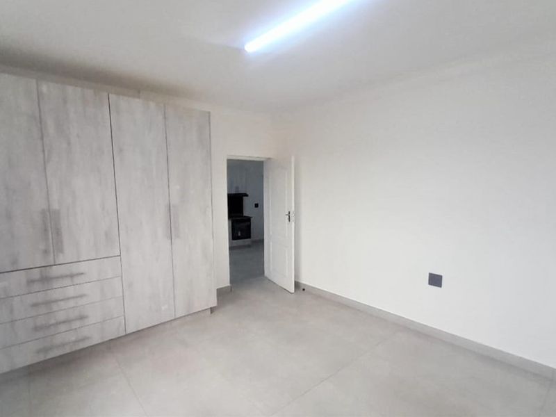 A clean, empty bedroom featuring modern light-wood built-in cupboards, neutral tiled flooring, and a simple interior door leading to an adjacent space.