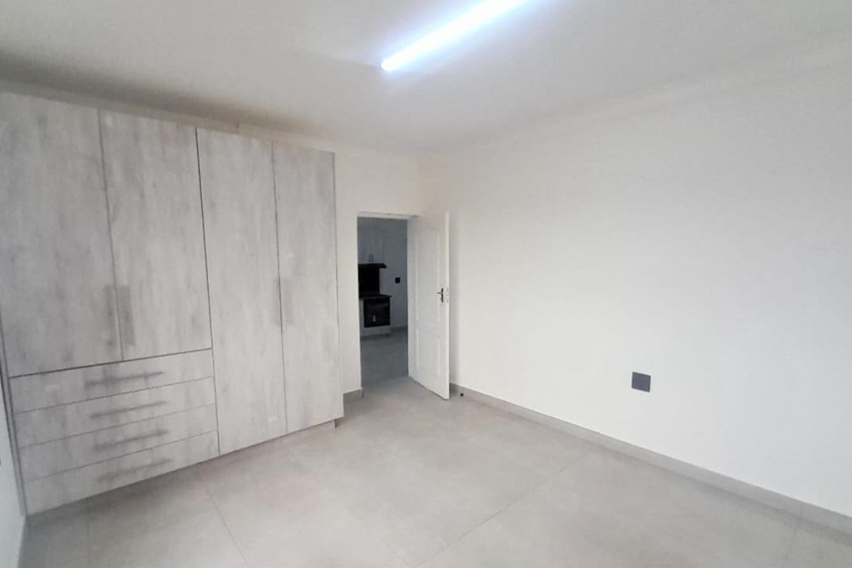 A clean, empty bedroom featuring modern light-wood built-in cupboards, neutral tiled flooring, and a simple interior door leading to an adjacent space.