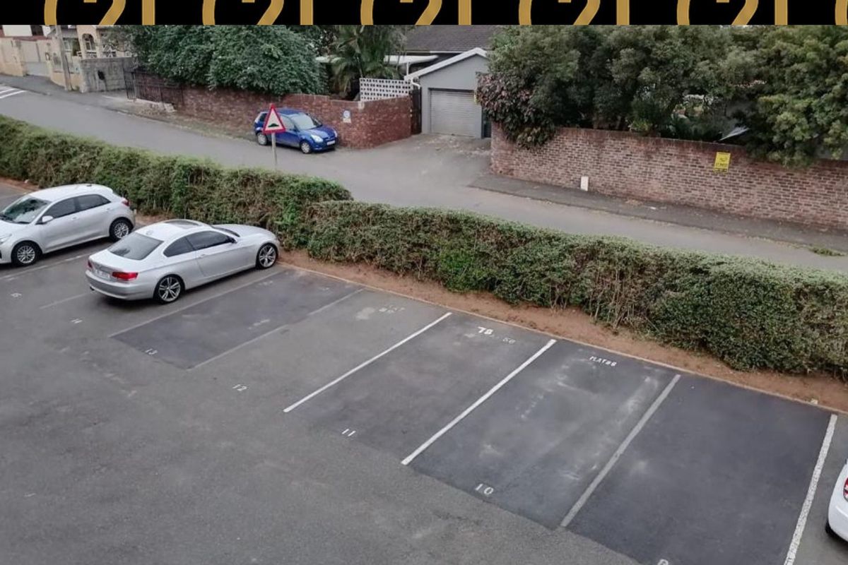 An elevated view of the apartment complex parking area, featuring marked asphalt bays and a perimeter brick boundary wall with established hedging.