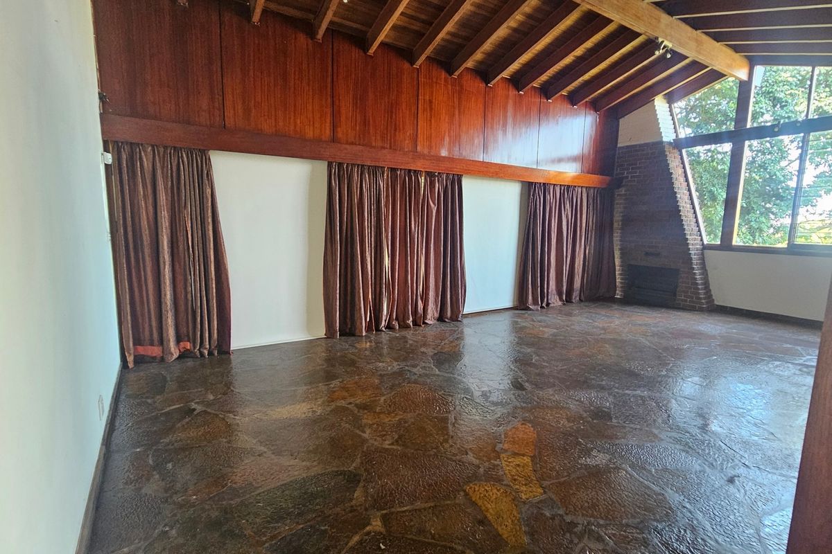 Spacious living room featuring exposed timber beam ceilings, unique stone flooring, a brick fireplace, and large windows providing ample natural light.