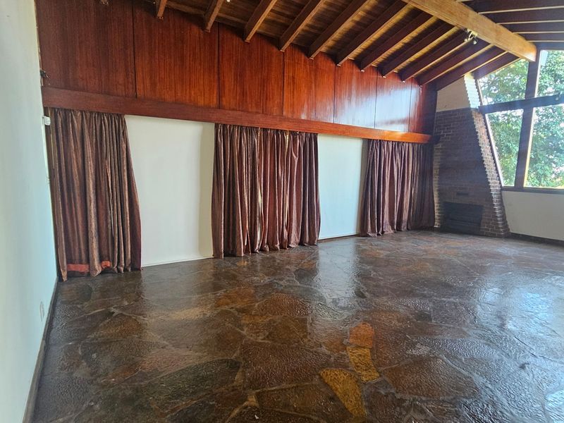 Spacious living room featuring exposed timber beam ceilings, unique stone flooring, a brick fireplace, and large windows providing ample natural light.