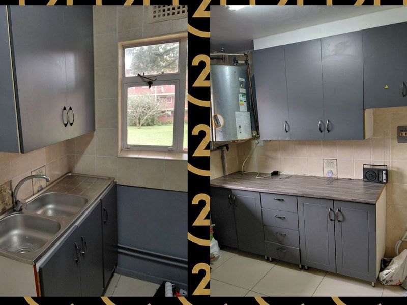 A compact kitchen featuring grey cabinetry, laminate countertops, and a double bowl stainless steel sink, requiring some maintenance and cosmetic updates.