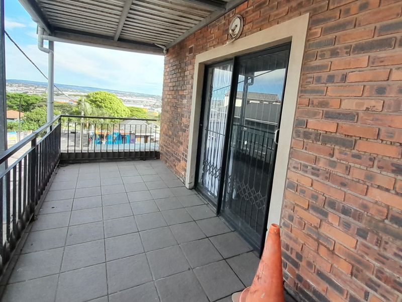 A covered balcony featuring tiled flooring, brick walls, and metal railings, offering an elevated view of the surrounding area.