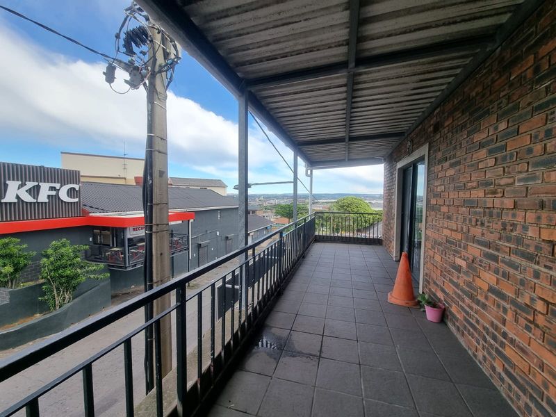 A covered balcony featuring tiled flooring, exposed brickwork, and metal railings, offering an elevated view of the surrounding urban area.