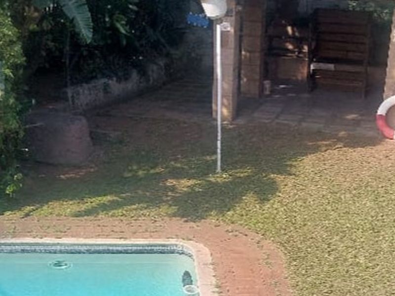 An outdoor pool area featuring a swimming pool with brick coping, a small covered patio space, and surrounding garden area.