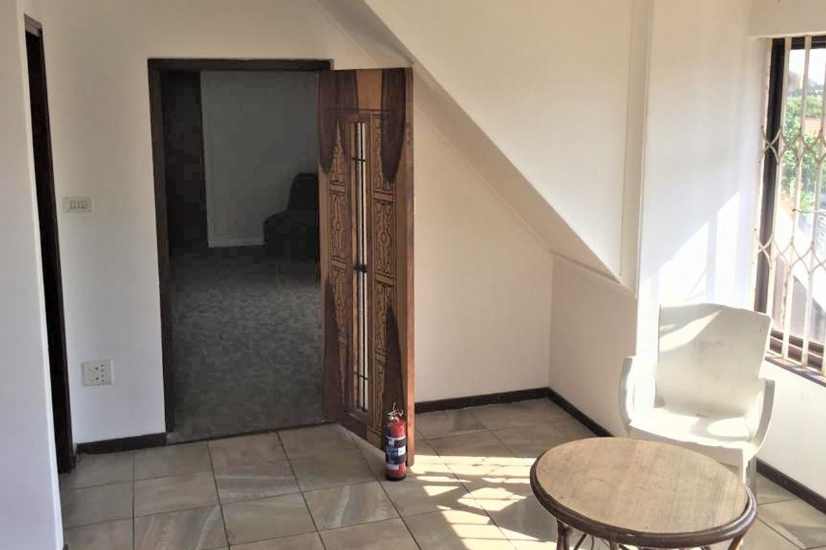 A sunlit living area featuring tiled flooring, a sloped ceiling, and a secure window with a metal gate, leading into an adjacent room.