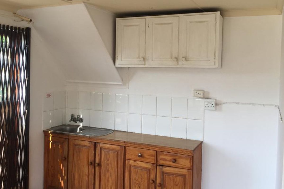 A compact kitchen area featuring wooden cabinetry, a stainless steel sink, and tiled splashback, currently in need of cosmetic updates.
