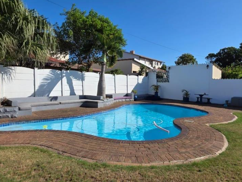 A well-maintained outdoor pool area featuring a kidney-shaped swimming pool with brick coping, surrounded by a lawn and secure boundary walls.