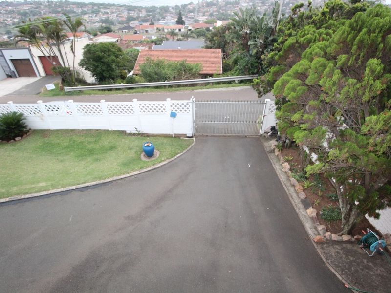 Elevated view of the property entrance featuring an asphalt driveway, metal security gate, and a decorative boundary wall with established garden landscaping.