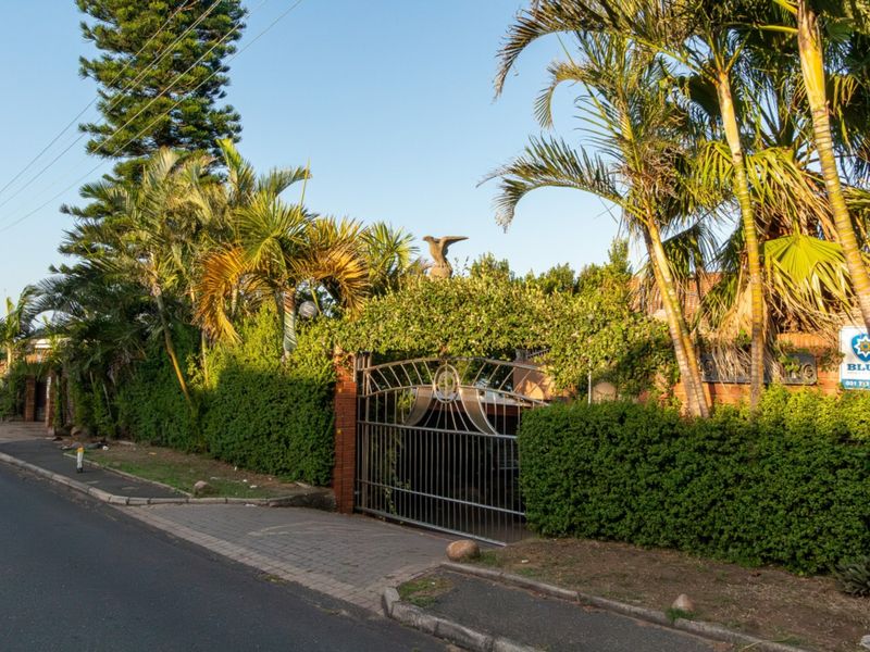 Street view of the property entrance featuring a metal security gate, brick boundary wall, and established garden hedging.
