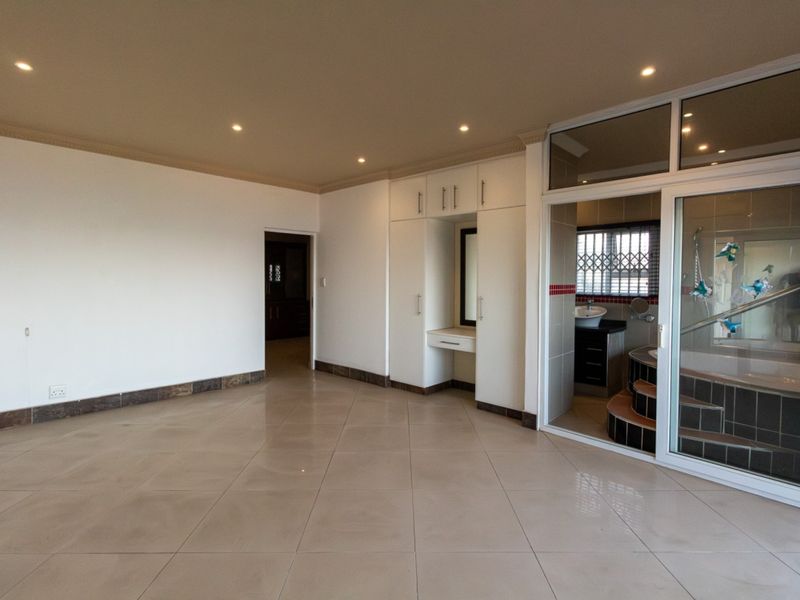 Spacious bedroom featuring neutral tiled flooring, built-in cupboards with a vanity area, and direct access to an en-suite bathroom through a sliding glass door.
