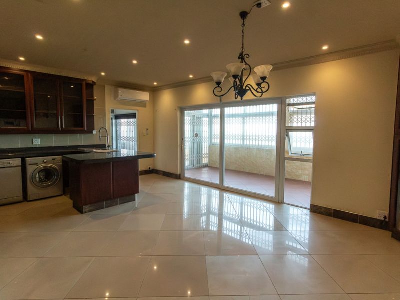 Spacious open-plan living area featuring tiled floors, recessed lighting, and a modern kitchen corner with granite countertops, leading to a covered balcony.