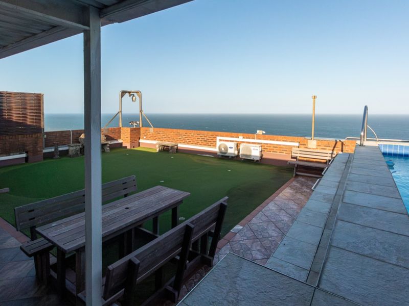 An elevated outdoor pool area featuring a swimming pool, artificial grass lawn, and a covered patio space with expansive ocean views.