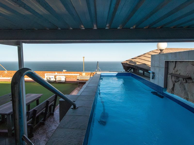 An elevated outdoor pool area featuring a lap pool, stone feature wall, and artificial turf, offering expansive ocean views from a covered patio space.