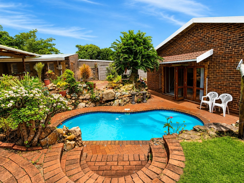 A well-maintained outdoor pool area featuring brick paving, landscaped rockery, and a covered patio adjacent to the main brick house structure.