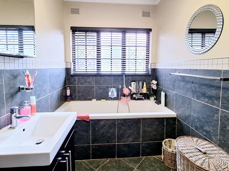A functional bathroom featuring a built-in bathtub, vanity basin, and dark tiled finishes with natural light provided by a window with venetian blinds.