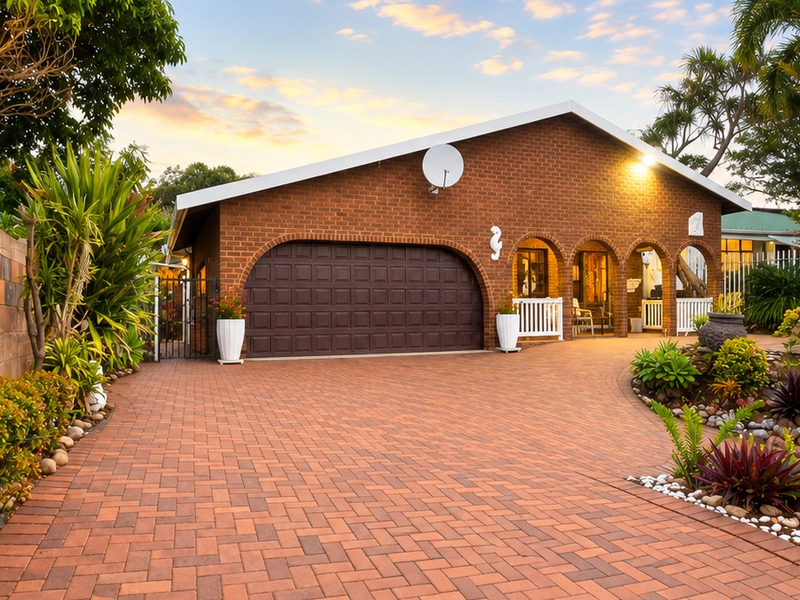 A well-maintained brick house featuring an arched garage door, a paved driveway, and landscaped garden beds, presenting a welcoming front facade.