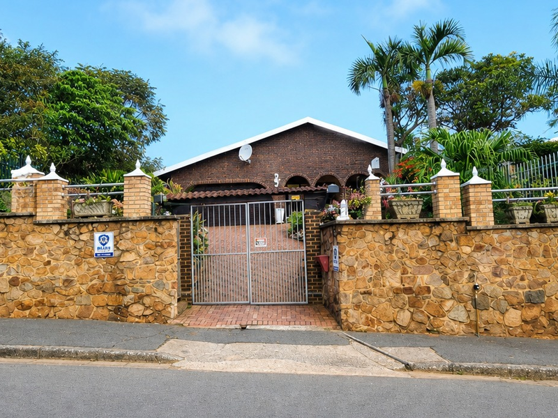 Front view of a residential property featuring a stone boundary wall, brick pillars, and a secure metal gate leading to a paved driveway.