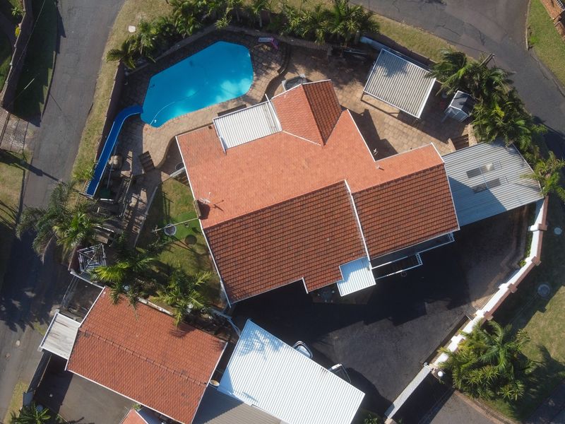 Aerial view of a residential property featuring a tiled roof house, swimming pool, paved driveway, and multiple carport structures on a corner lot.