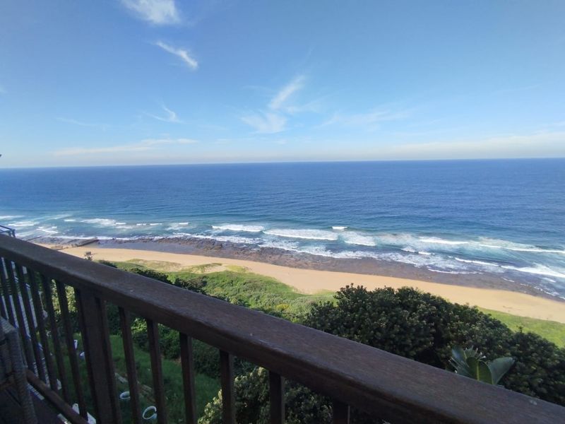 A scenic elevated view overlooking the coastline, sandy beach, and ocean, framed by a timber balcony railing.