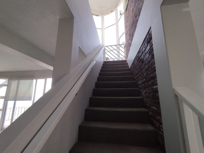 A staircase featuring an exposed brick wall, white handrails, and a large window providing natural light, leading to an upper level with a safety gate.