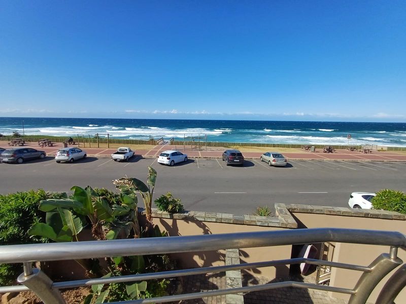 Elevated coastal view overlooking a public parking area, promenade, and the ocean, captured from a private balcony with a stainless steel balustrade.