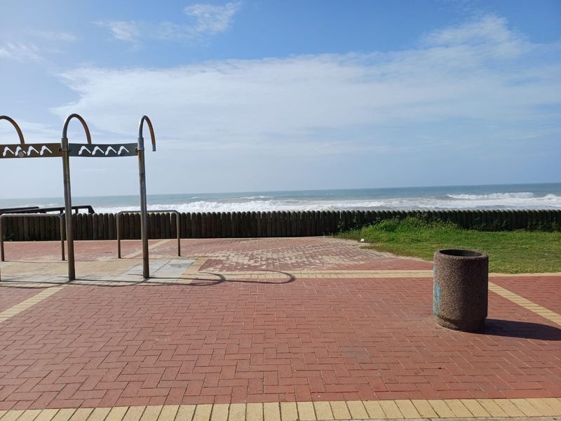 A scenic outdoor area featuring a paved walkway, an outdoor shower structure, and a timber retaining wall overlooking the ocean.