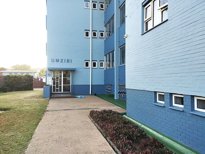 Exterior view of the Umzibi apartment building entrance, featuring a blue brick facade, paved walkway, and landscaped garden beds.
