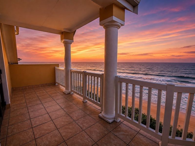 A spacious covered balcony featuring tiled flooring and white balustrades, offering stunning direct ocean and beach views at sunset.