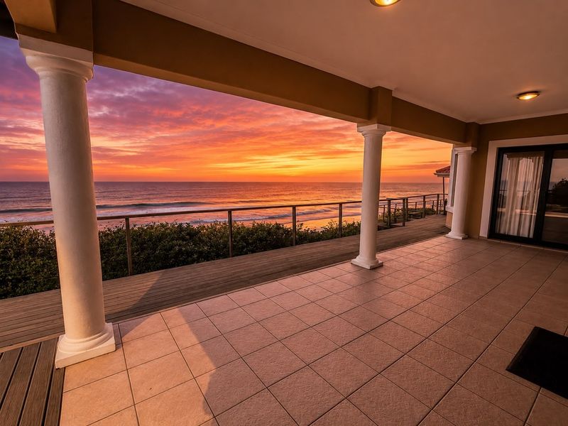 Spacious covered patio featuring tiled flooring, classical columns, and a glass balustrade, offering stunning elevated ocean views at sunset.