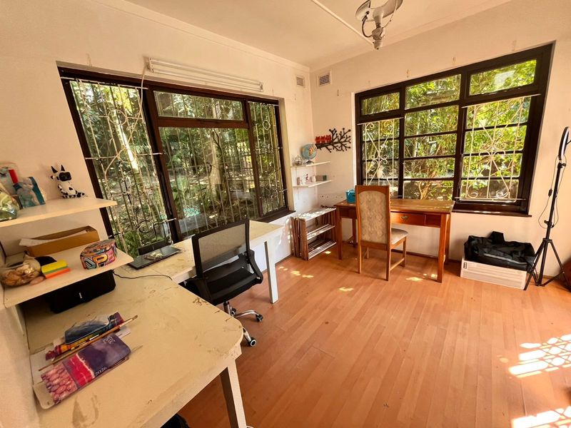 A sunlit home office featuring laminate flooring and large windows with security bars, offering a bright workspace with garden views.
