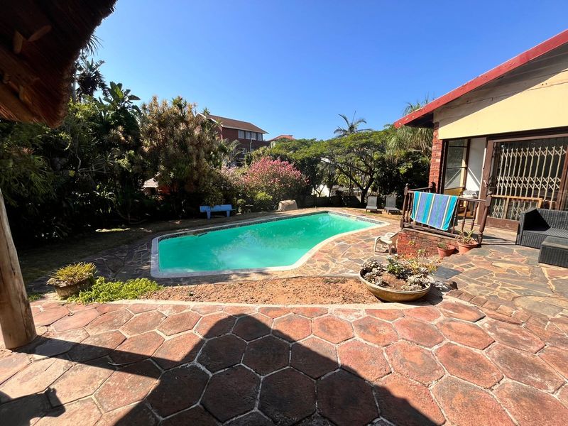 Outdoor entertainment area featuring a swimming pool, stone paved patio, and a thatched roof structure set within a landscaped garden.