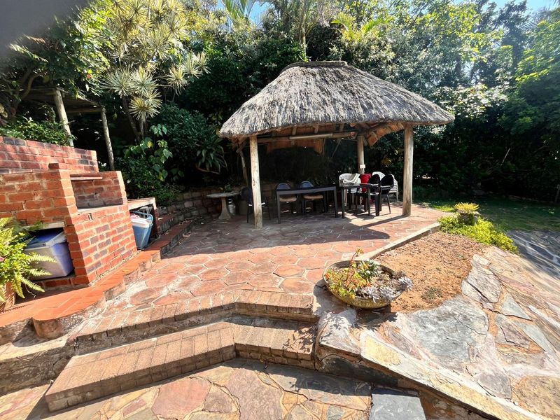 Outdoor entertainment area featuring a traditional thatched lapa, brick built-in braai, and stone paved patio set within a lush garden.