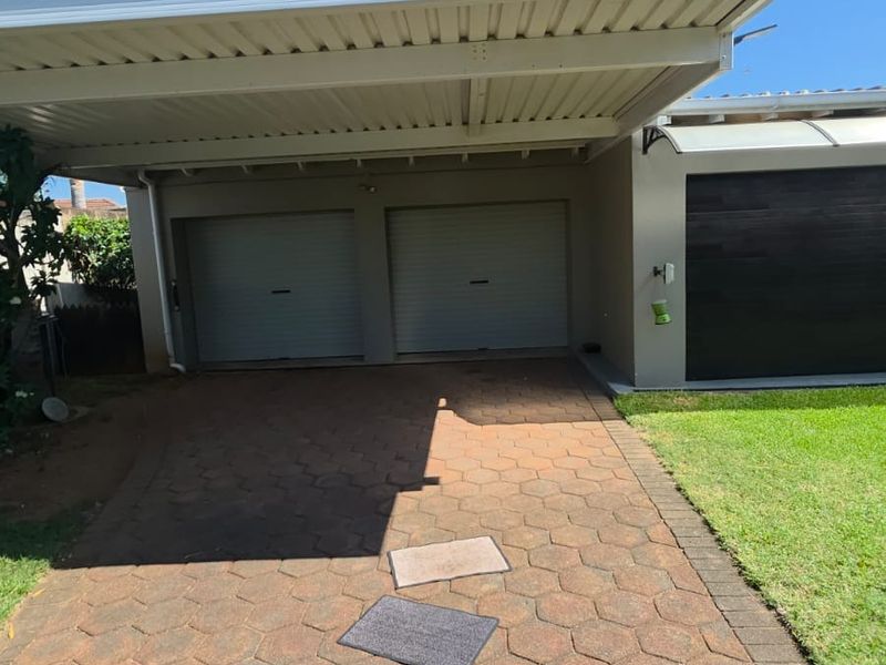 A well-maintained front exterior featuring a double garage with roller shutter doors, a covered carport, and a paved driveway.