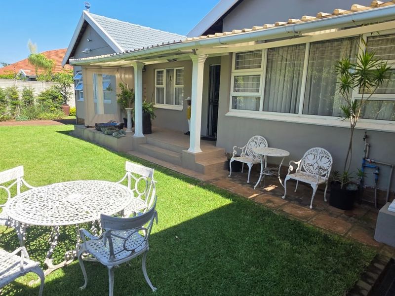 A well-maintained rear exterior view featuring a covered patio area, tiled flooring, and a spacious green lawn with boundary walling.