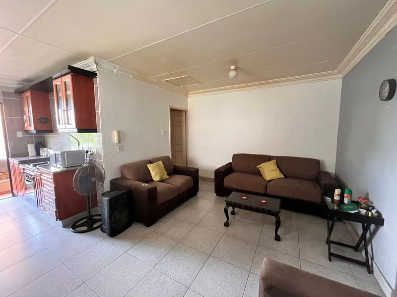 Open-plan living area featuring tiled flooring and an adjoining kitchen with built-in cabinetry, offering a functional layout in need of cosmetic updates.