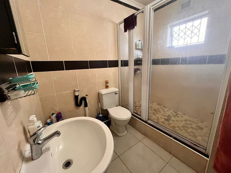A functional bathroom featuring a shower enclosure, pedestal basin, and toilet with neutral wall tiling and a security-grille window.