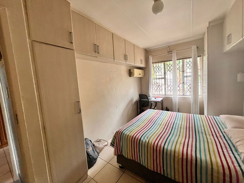 A bedroom featuring built-in cupboards, tiled flooring, and an air conditioning unit, with security bars installed on the window.